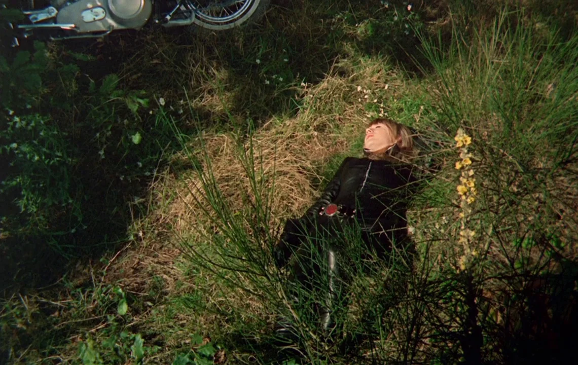 The Girl on a Motorcycle (1968) movie still: wide — A woman in a black leather suit lies prone in overgrown, patchy meadow grass. The partial wheel…