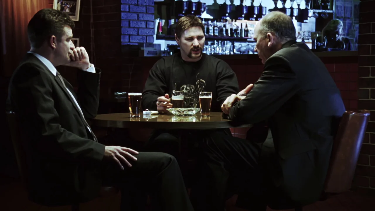 Chopper (2000) movie still: medium — A dimly lit pub interior features dark brick walls and a back-lit bar area. Three men sit at a round table with…