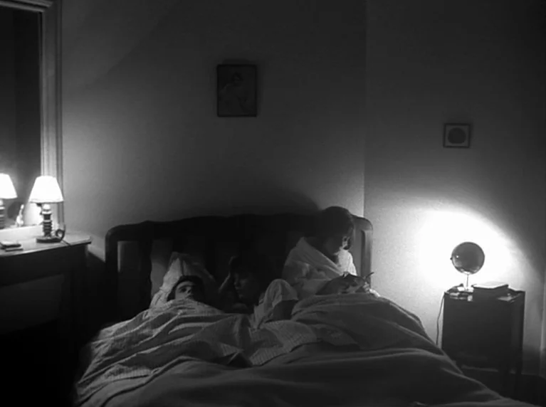 Masculin Féminin (1966) movie still: medium — A dimly lit room at night with two people lying in a double bed. A small lamp on a nightstand…