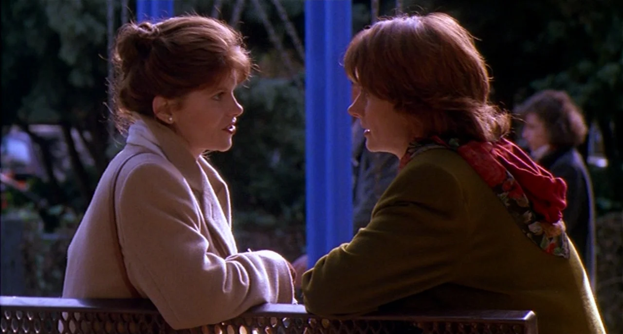 Raising Cain movie still: medium — Two women sit on a dark metal bench in a park during the daytime. The women are engaged in a conversation while…