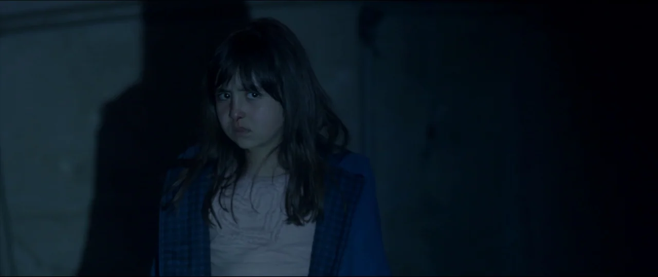 Under The Shadow (2016) movie still: medium — A girl stands centered in a dark, nondescript interior space with minimal architectural detail. She…