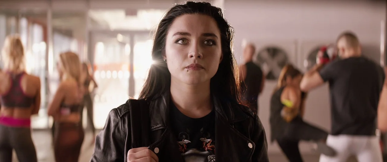 Fighting With My Family (2019) movie still: closeup — A fitness center interior has blurry people working out in the background under bright light.…