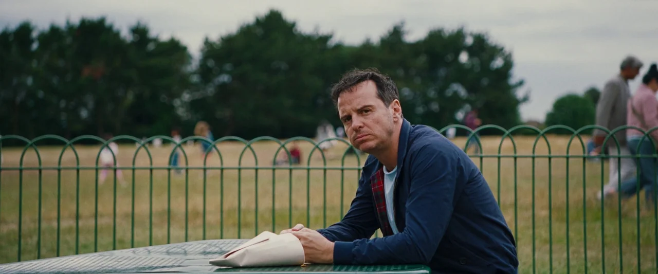 All Of Us Strangers (2023) movie still: medium — A green metal table sits in an open public space with a low fence. A man sits at the table in a…