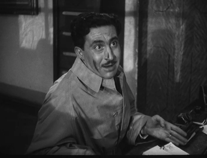 The White Sheik (1952) movie still: medium — A cluttered room features a wooden cabinet and a desk with scattered papers. A man in a trench coat…