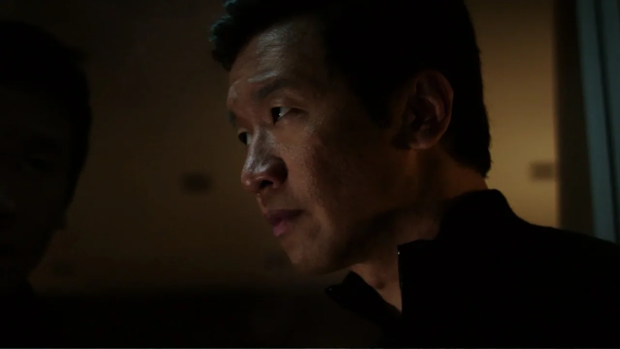 Skyscraper (2018) movie still: closeup — A dimly lit room with heavy shadows obscuring the background. An East Asian man looks toward the side of…