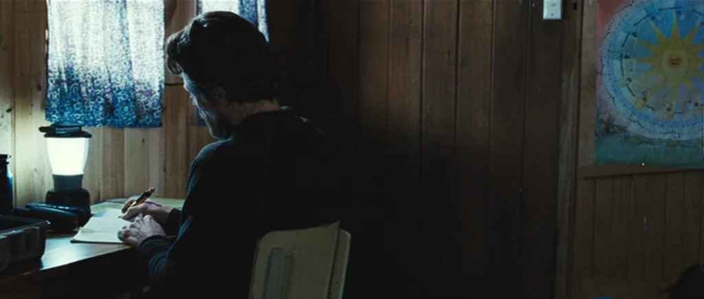 The Hunter (2011) movie still: medium — A rustic cabin room with vertical wood paneling and a colorful circular sun poster. A man sits at a desk…
