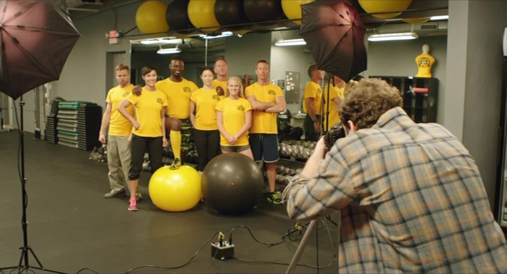 Results (2015) movie still: medium — A group of six people stand in a line wearing matching yellow team shirts. A photographer stands in the…