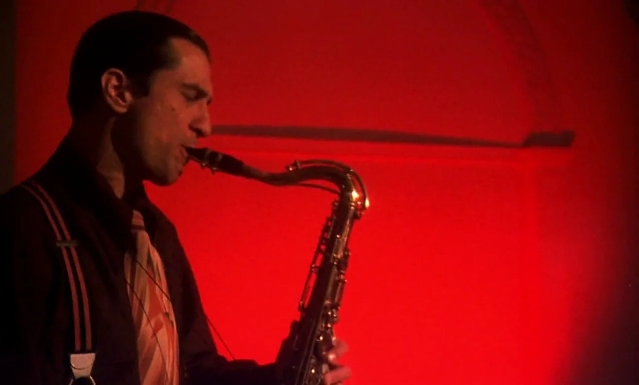 New York, New York (1977) movie still: medium — A man plays a saxophone in front of a flat, solid red wall surface. He wears a dress shirt and…