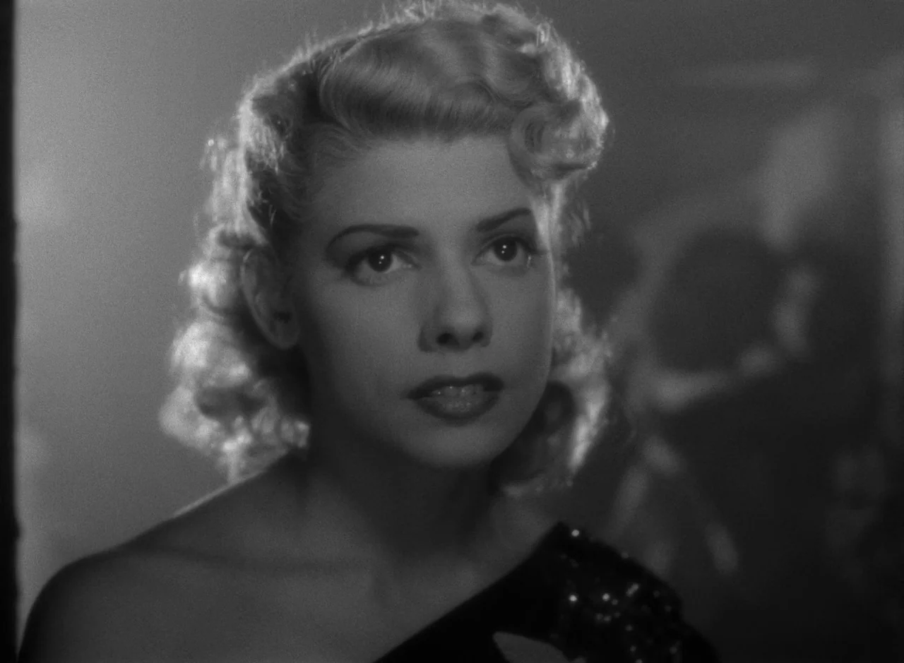 Victims of Sin (1951) movie still: closeup — A dimly lit indoor space with a dark background and high-contrast lighting. A young woman with blonde…
