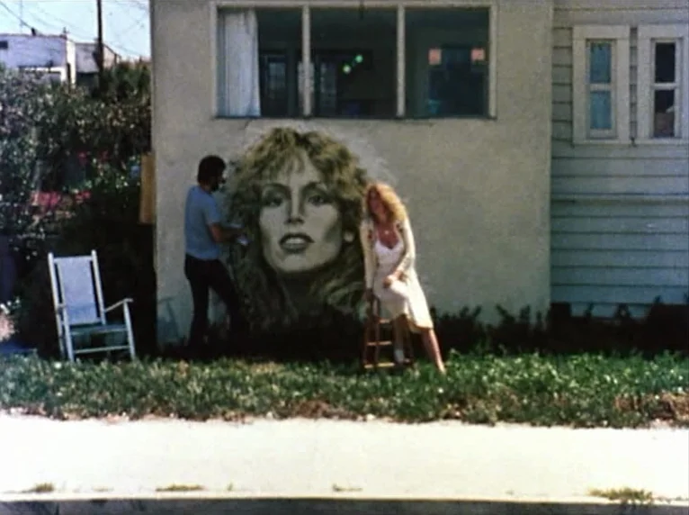 Mur Murs (1981) movie still: wide — A man paints a large portrait mural on the side of a beige residential house. A blonde woman sits on a small…