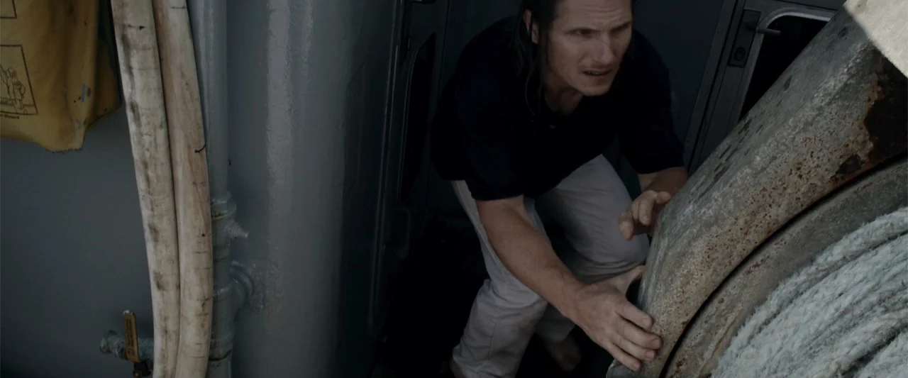 Mermaid Down (2019) movie still: medium — A cramped and weathered boat deck contains metal structures and heavy maritime ropes. A man in a dark…