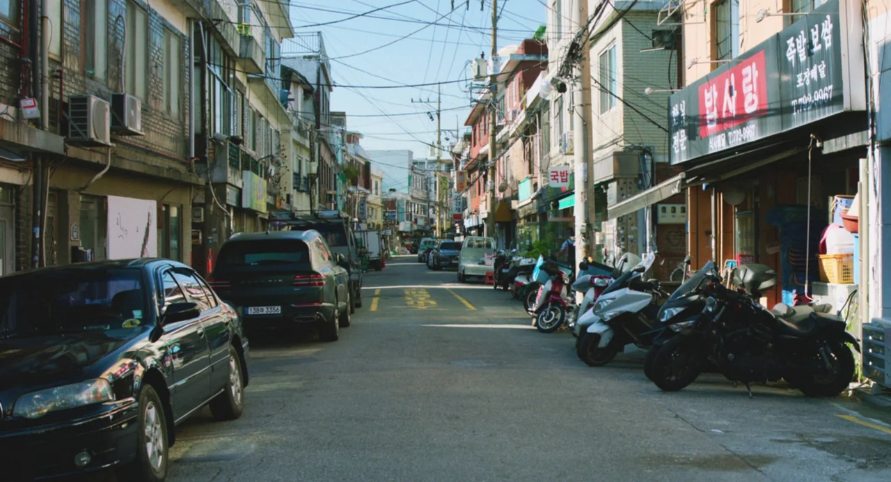 Return To Seoul (2022) movie still: establishing — A narrow urban road in South Korea is flanked by low-rise commercial buildings. Several cars and…
