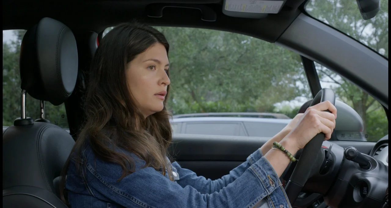 One True Loves (2023) movie still: medium — A woman sits in the driver seat of a car during the day. She grips the steering wheel with both hands…