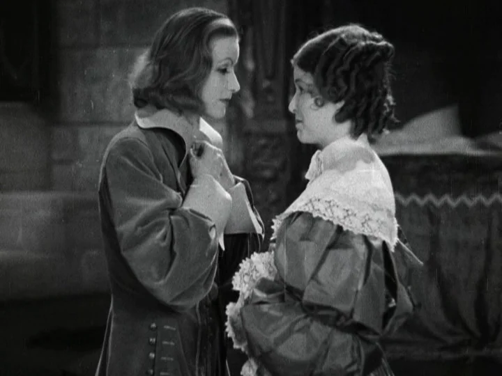 Queen Christina (1933) movie still: medium — An opulent, dim chamber features stone architecture and shadows in the background. Two women in period…
