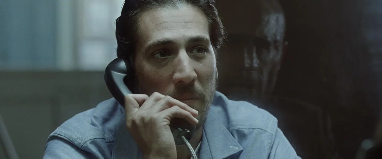 Anna (2013) movie still: closeup — A man sits before a glass partition in a sterile visitation room setting. The man holds a black telephone…