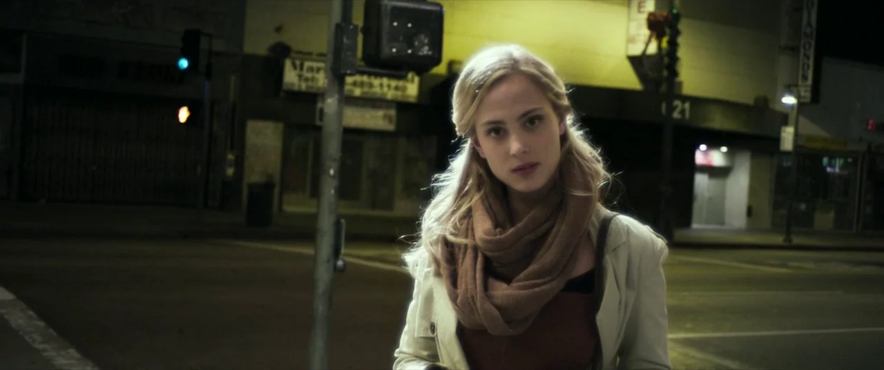 Maniac (2012) movie still: medium — An empty urban street intersection at night with dim yellow lighting and dark buildings. A young woman in a…