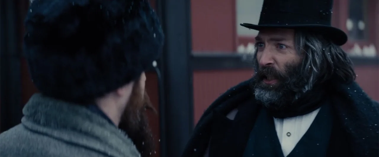 I Heard the Bells (2022) movie still: medium — Two men stand outdoors near a building exterior during a light snowfall. The man in the foreground…
