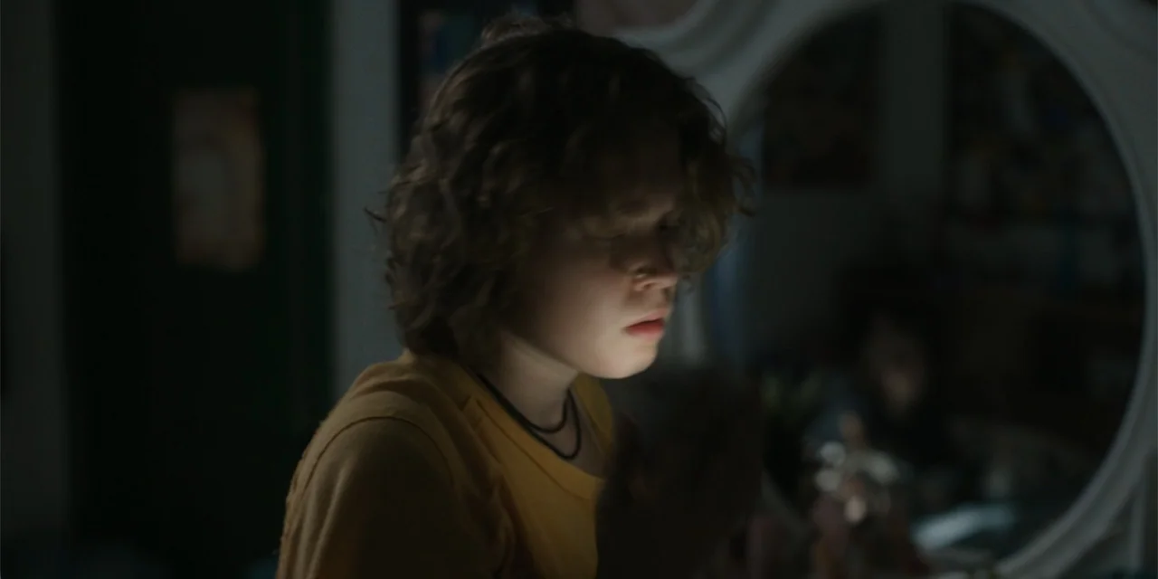 Caddo Lake (2024) movie still: medium — A dark bedroom interior features a rounded mirror and blurred vanity furniture in back. A teenager stands…