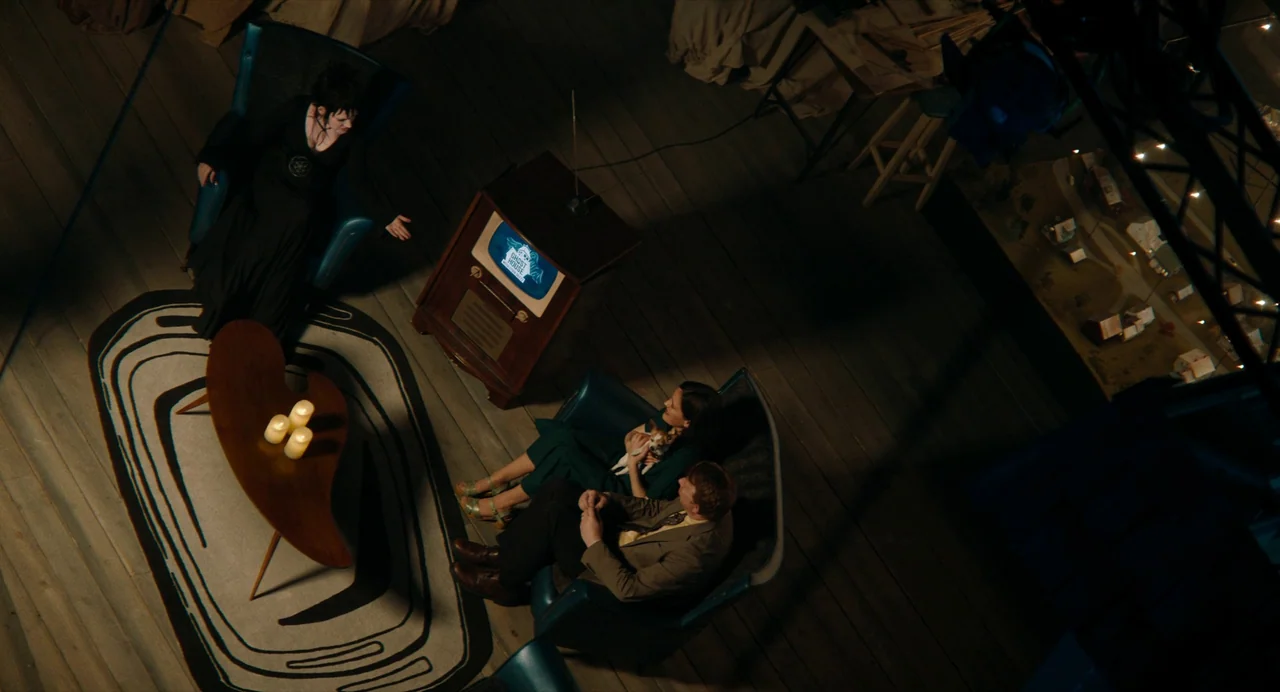Beetlejuice Beetlejuice (2024) movie still: wide — A dark, wood-floored living room features mid-century modern armchairs arranged around a central…