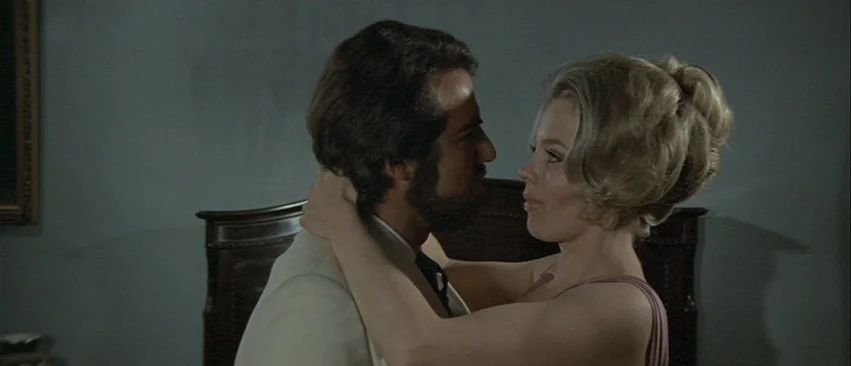 Whity (1971) movie still: medium — A dark bedroom features a large dark wooden headboard against a plain grey wall. A man and a woman face each…