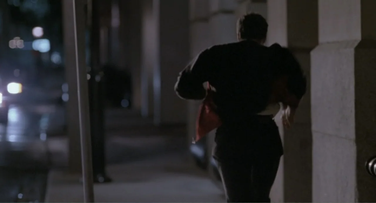 F/X2 (1991) movie still: medium — A person walks along a concrete sidewalk beside building pillars at night. The subject moves away from the camera…