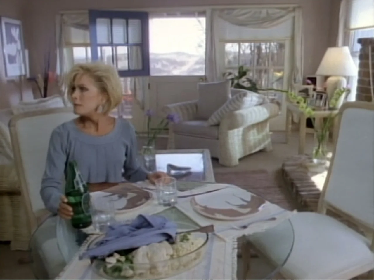 My First Love (1988) movie still: medium — A light-filled room features a glass dining table set for a meal. A blonde woman sits at the table…