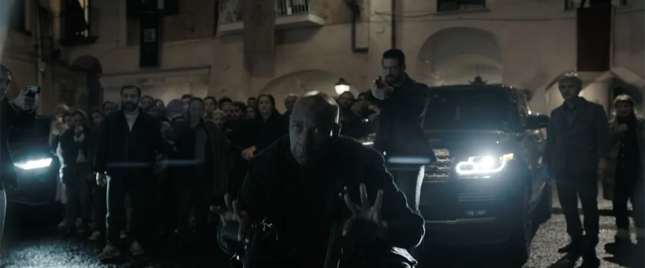The Equalizer 3 (2023) movie still: medium — An outdoor town square features ancient stone architecture illuminated at night by vehicle headlights.…