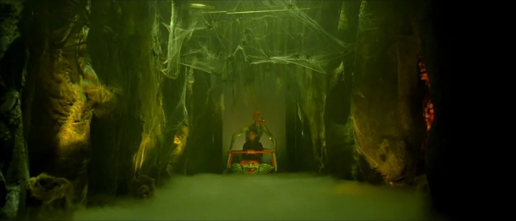 Amelie (2001) movie still: wide — A dark ride tunnel features thick green fog, hanging spiderwebs, and textured rock-like walls. A person sits in a…