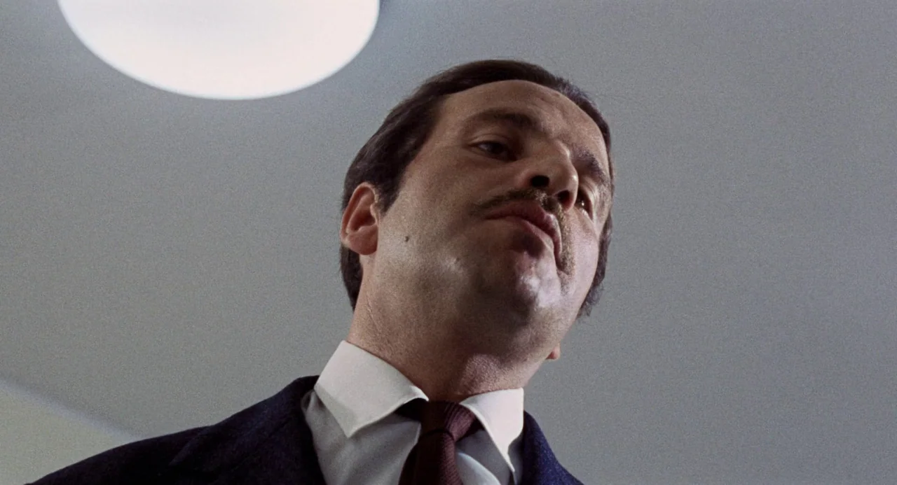 Investigation of a Citizen Above Suspicion (1970) movie still: closeup — A plain interior room features a stark white wall with a round light…