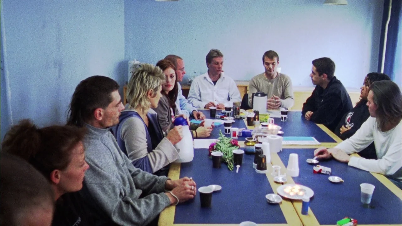 Pusher III (2004) movie still: wide — A group of people sit around a large rectangular table in a room with blue walls. The table is cluttered with…