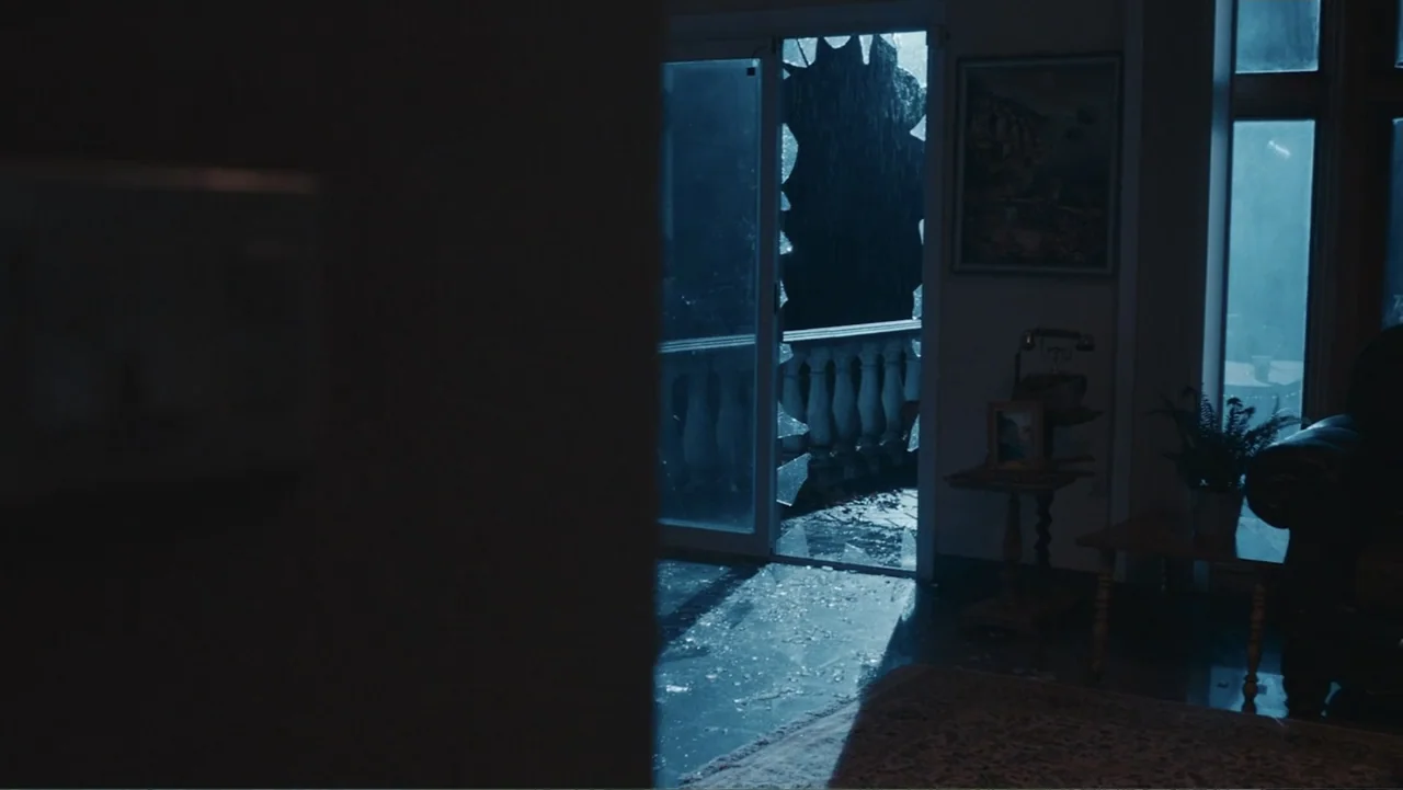 Terrifier 2 (2022) movie still: wide — A dark living room features a shattered glass door leading out to a balcony. Rain falls outside the broken…