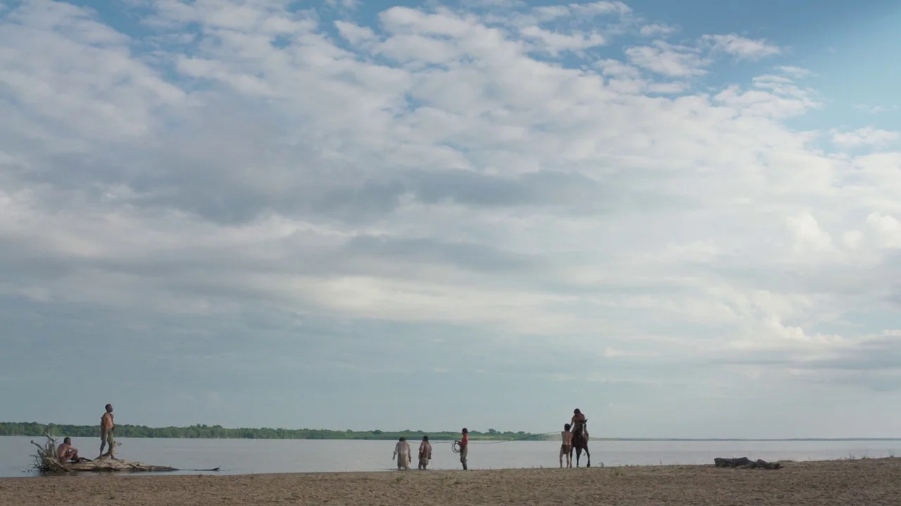 Zama (2017) movie still: wide — A wide sandy riverbank sits under a large cloudy sky near a vast river. A group of people stand along the shore…