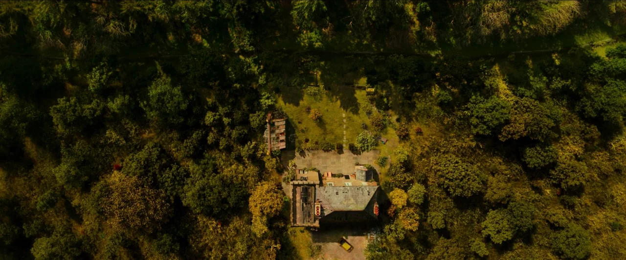 Unwelcome (2022) movie still: establishing — A secluded house sits in a clearing surrounded by dense, overgrown woods. The building has a stone…