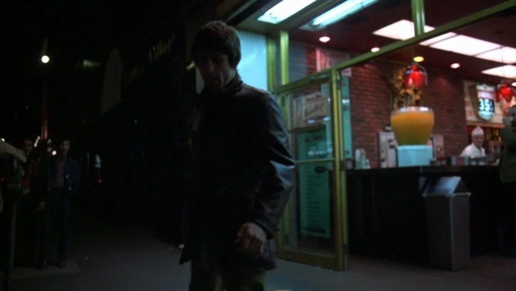 Mean Streets (1973) movie still: medium — A man walks along a dark city sidewalk at night past a luminous shop entrance. A large, amber-colored…