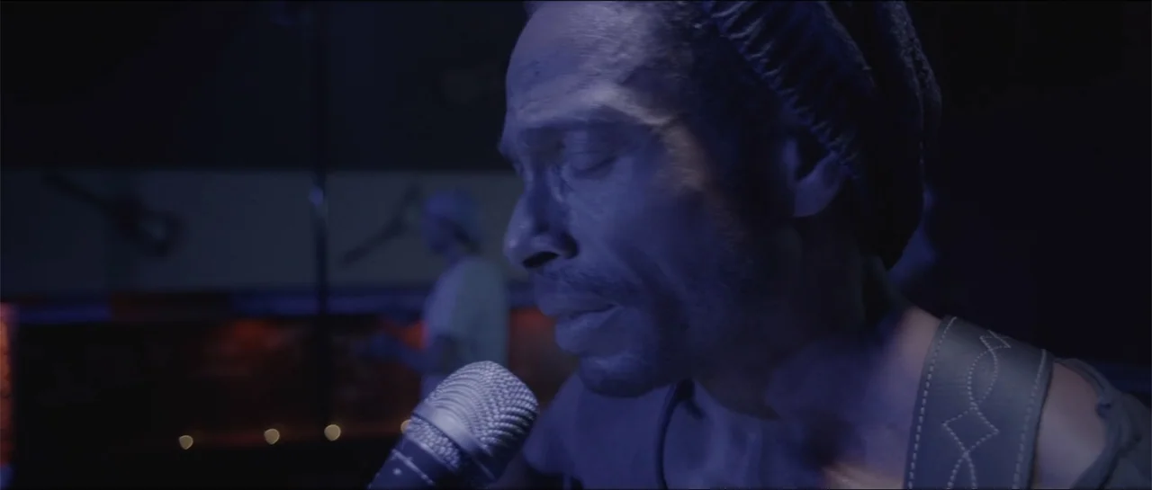 All She Wrote (2018) movie still: closeup — A dark music venue features a man singing into a microphone on stage. The singer stands in profile while…