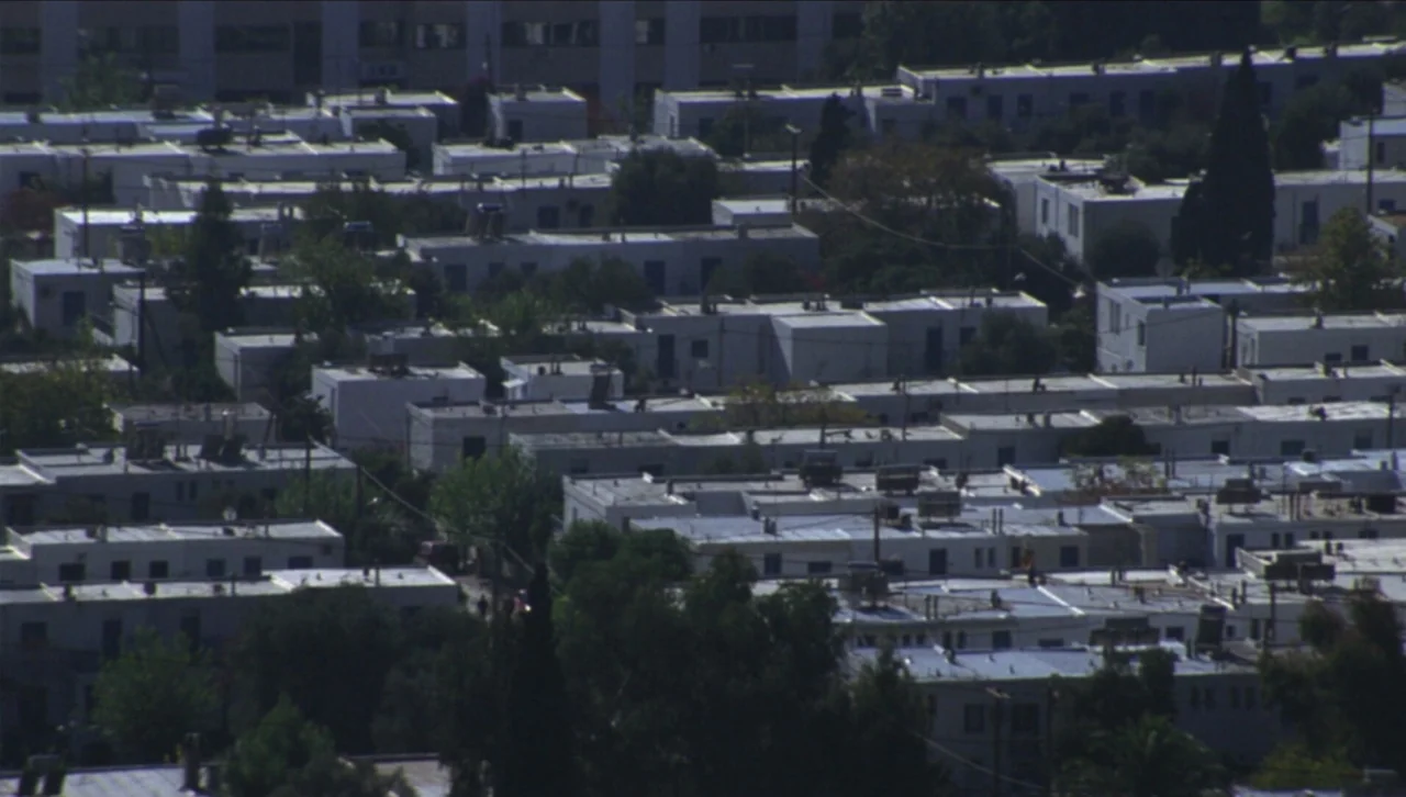 Attenberg (2010) movie still: establishing — A dense residential neighborhood features rows of flat-roofed white buildings and green trees. The…
