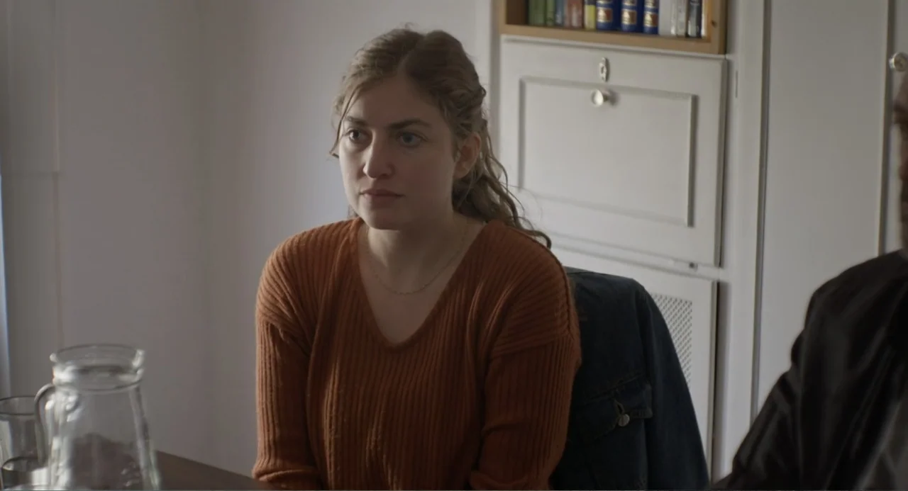 An Odd Turn (2024) movie still: medium — A light-walled room features a white cabinet and a dark table in the foreground. A woman in a rust-colored…