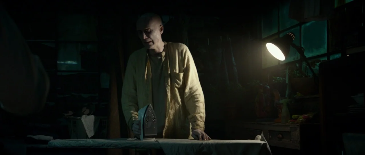 Logan (2017) movie still: medium — A dark, cluttered workspace is lit by a small directional lamp on a table. An elderly man with a shaved head…