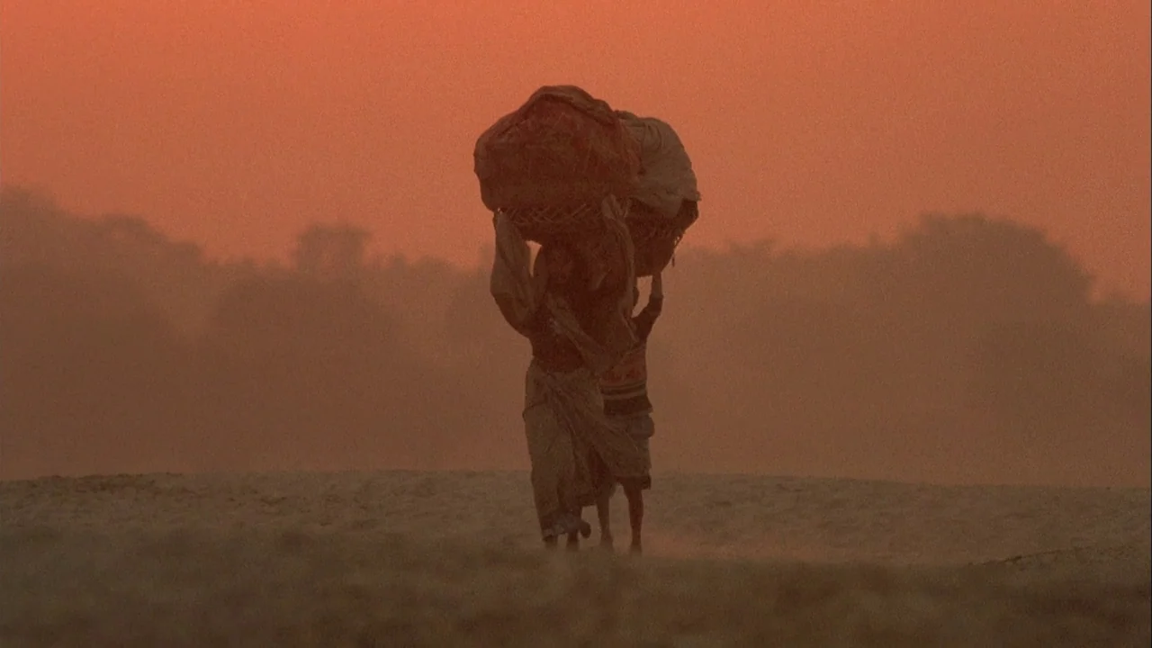 Powaqqatsi (1988) movie still: fullbody — A vast sandy landscape stretches under a hazy orange sunset sky. Two figures walk forward while balancing…
