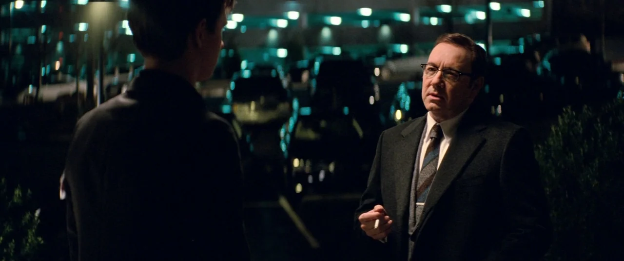Baby Driver (2017) movie still: medium — An outdoor parking lot at night with rows of parked vehicles in the background. A man in a suit speaks to…