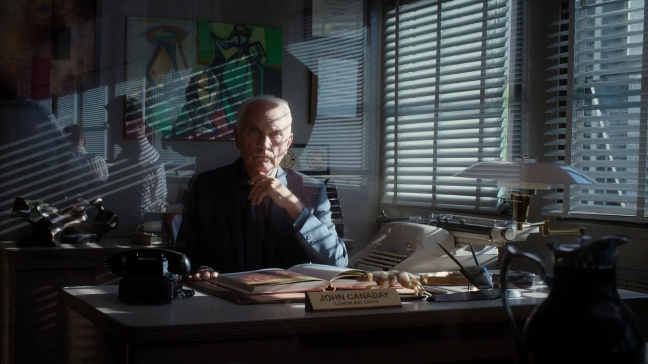 Big Eyes (2014) movie still: medium — An office with venetian blinds, a typewriter, and a nameplate reading John Canaday. The elderly man rests his…
