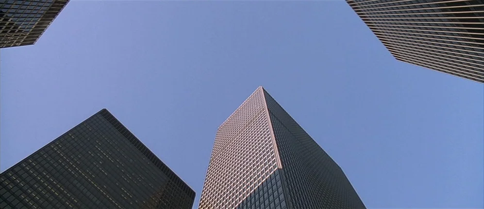 American Psycho (2000) movie still: establishing — Four tall glass skyscrapers surround an open section of bright clear blue sky. The geometric…