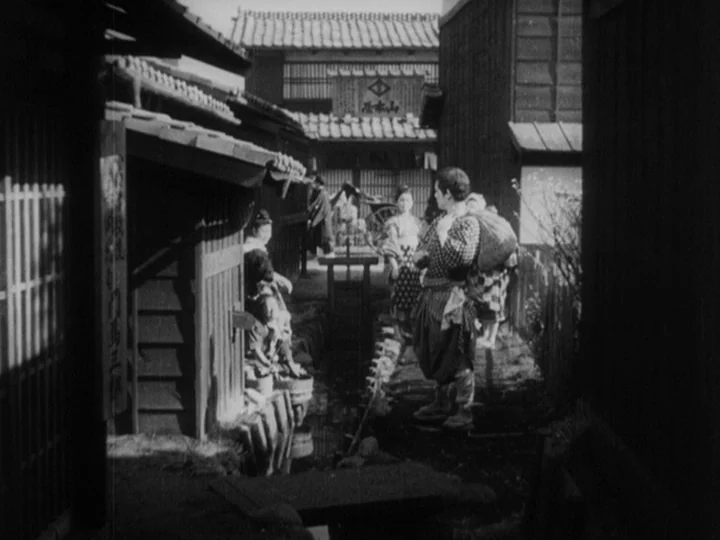 Sanshiro Sugata (1943) movie still: fullbody — A narrow Japanese neighborhood alleyway features wooden buildings with tiled roofs and a ditch. A man…