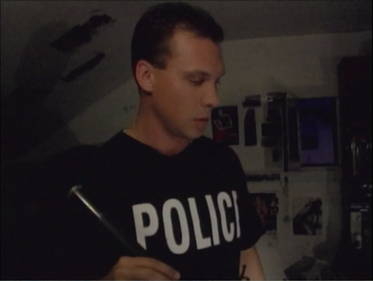 Addiction (2024) movie still: medium — A dark indoor setting with posters visible on the back wall. A man in a black police t-shirt holds a baton…