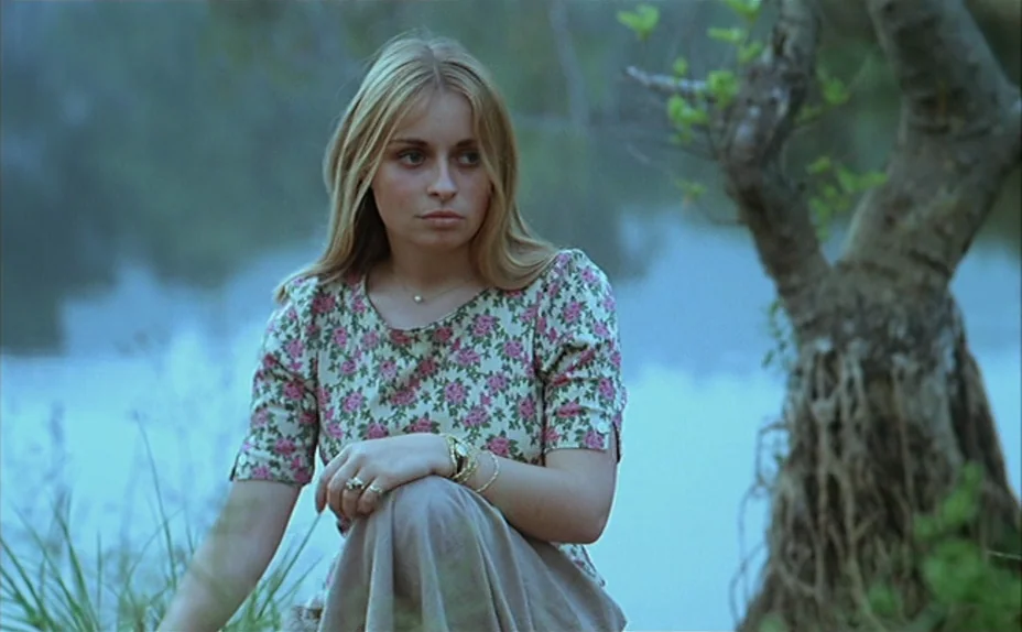 L’une Chante, l’Autre Pas (1977) movie still: medium — A woman crouches beside a gnarled tree on a lush green riverbank. The river flows in the…