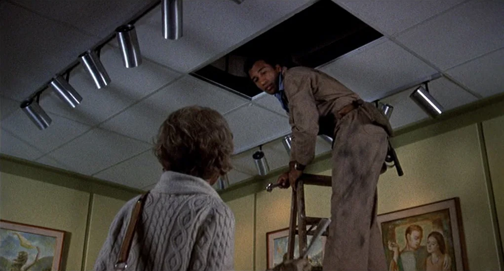 Family Plot (1976) movie still: medium — An interior office space features beige paneled walls adorned with framed art prints. A man climbs a wooden…