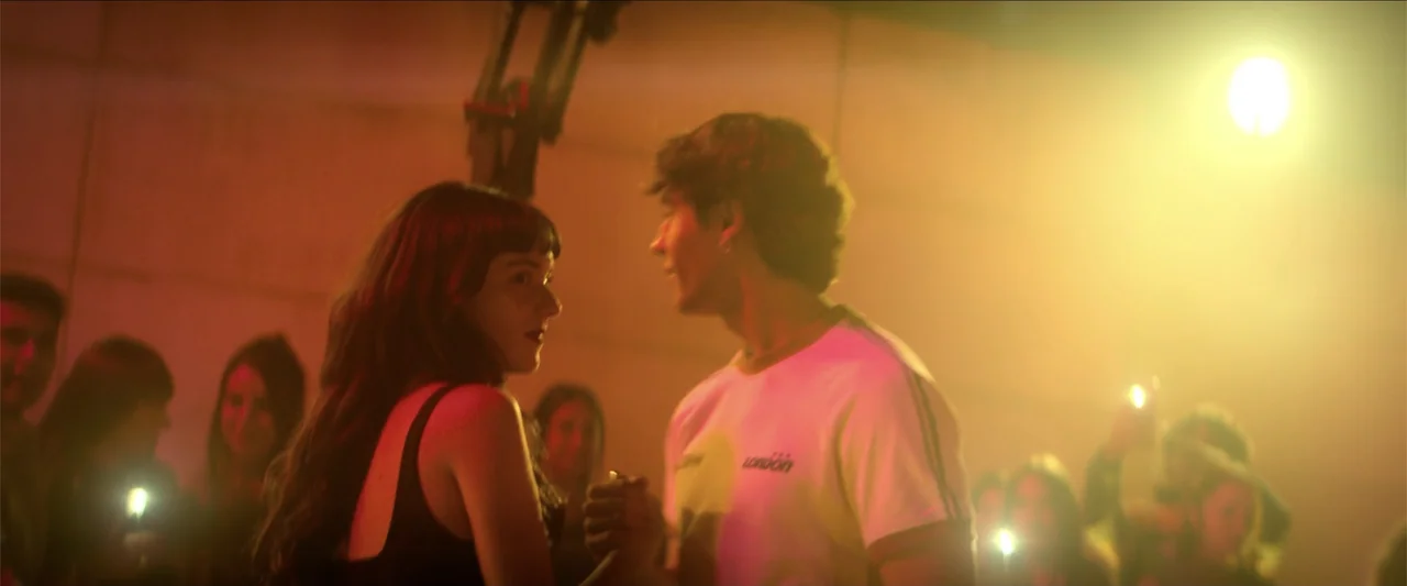 Tell Me Softly (2025) movie still: medium — A crowded indoor venue features walls lit by harsh golden stage lighting and shadows. A young man and…