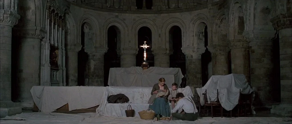 Jude (1996) movie still: wide — A stone Romanesque interior with arched columns and a central altar draped in cloths. A family sits on the floor…