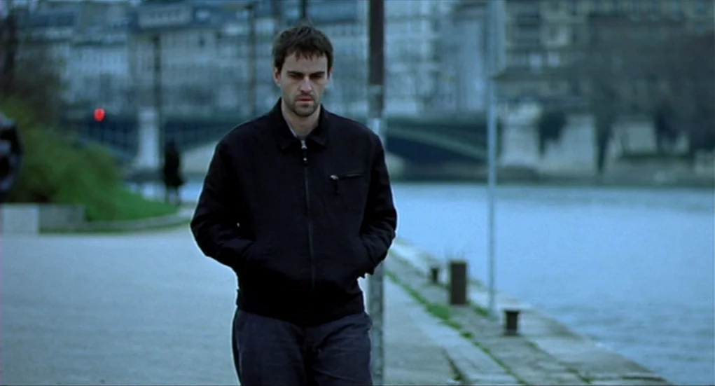 Tiresia (2003) movie still: medium — A riverside walkway in an urban area with a stone embankment and distant bridge. A man walks directly toward…
