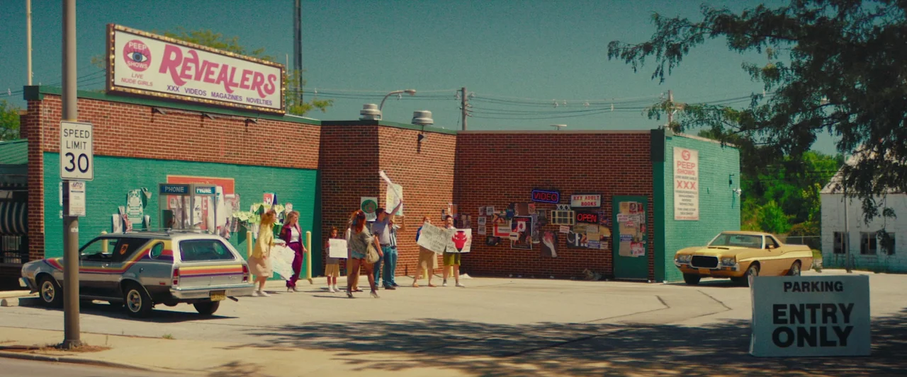 Revealer (2022) movie still: wide — A brick storefront for a peep show venue features neon signage and posters. A small group of people stand in the…